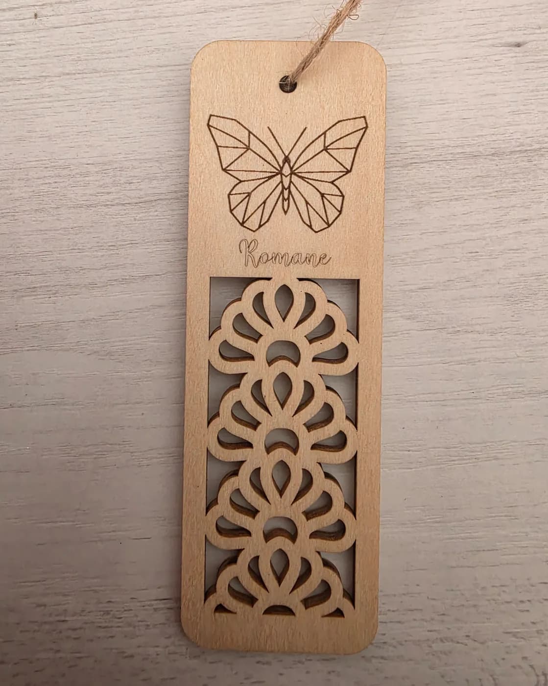 Marque-page en bois clair avec une ficelle de jute : un papillon gravé en haut, un prénom en écriture et le bas découpé en dentelle ajourée.